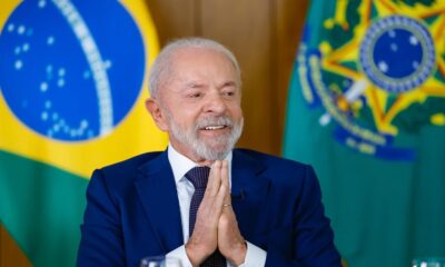 Lula sorrindo, de terno azul, com mãos unidas e bandeiras do Brasil