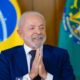 Lula sorrindo, de terno azul, com mãos unidas e bandeiras do Brasil