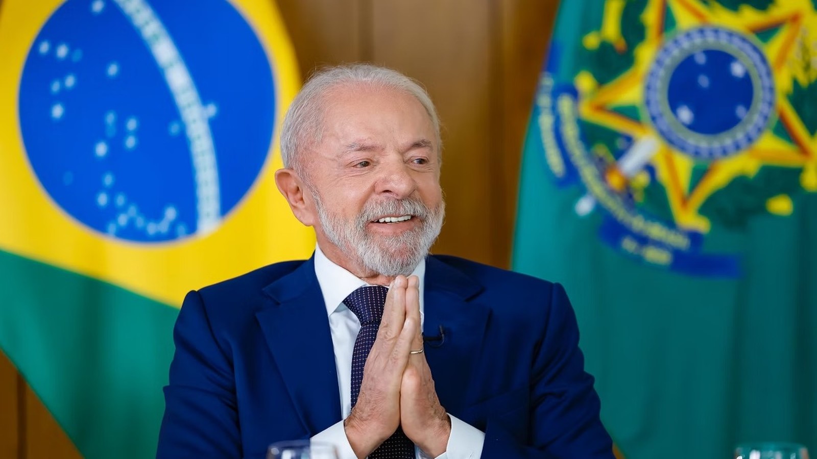Lula sorrindo, de terno azul, com mãos unidas e bandeiras do Brasil