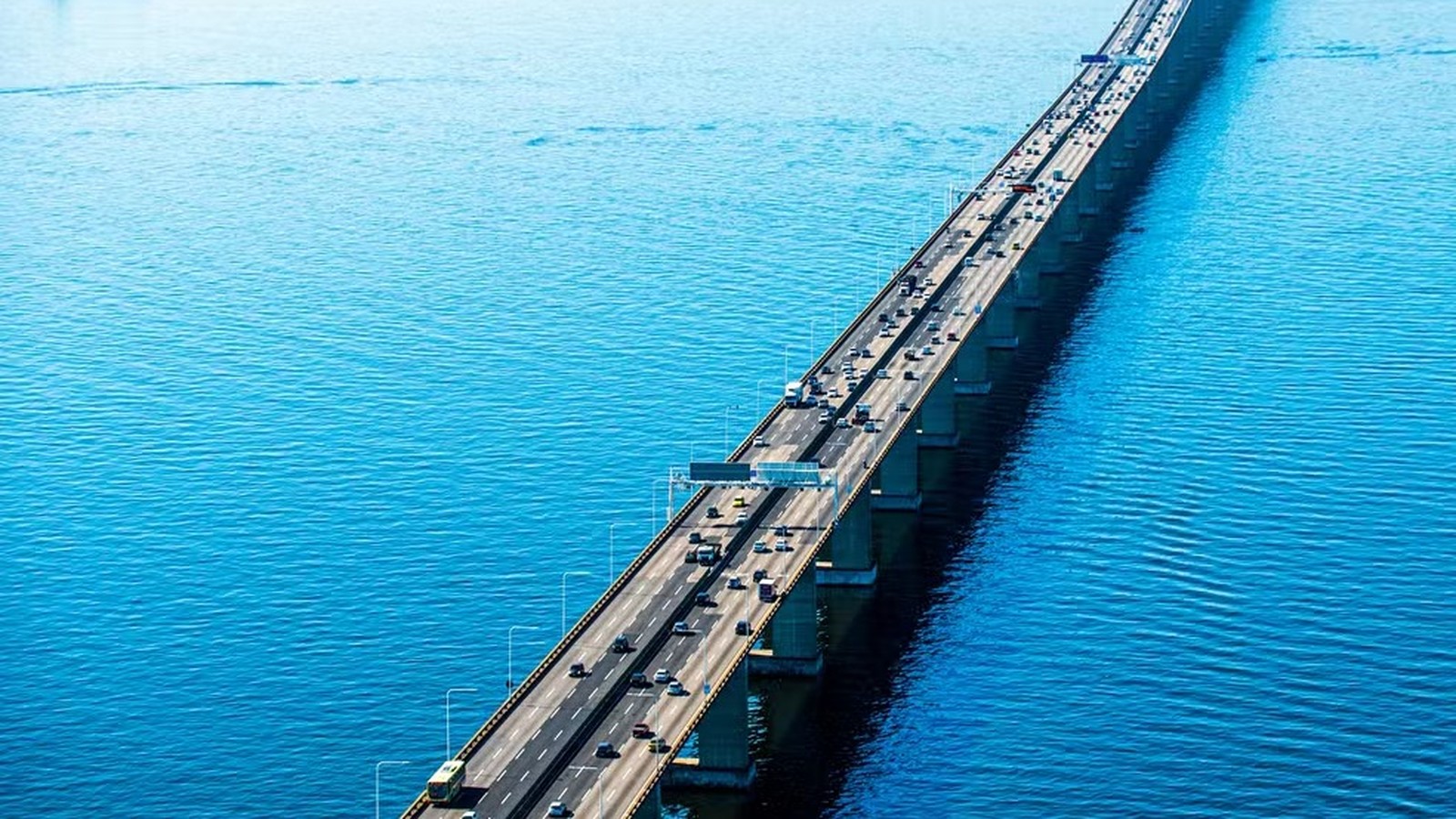 Vista aérea da Ponte Rio-Niterói sobre a água azul, com muitos carros e caminhões