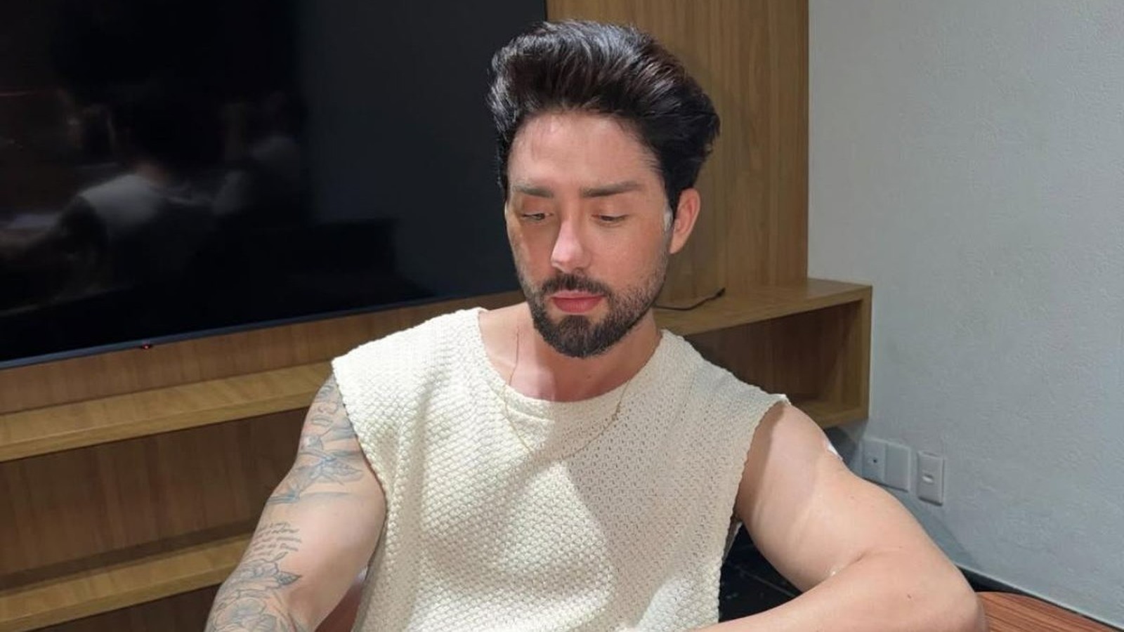 Rico Melquiades, homem barbudo, camiseta creme sem manga, olhando para baixo