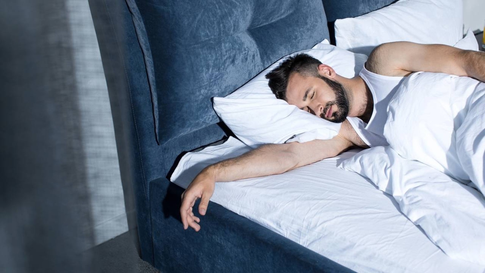 Homem barbudo dormindo profundamente na cama com travesseiro e lençóis brancos
