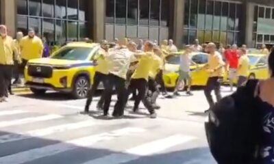 Homens em camisas amarelas brigam em rua com taxis amarelos ao fundo no aeroporto