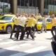 Homens em camisas amarelas brigam em rua com taxis amarelos ao fundo no aeroporto