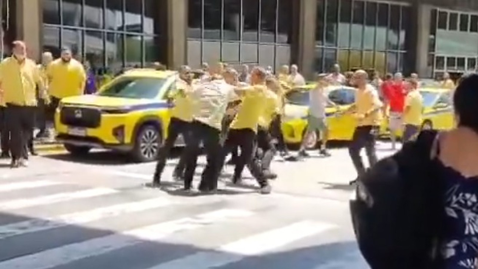 Homens em camisas amarelas brigam em rua com taxis amarelos ao fundo no aeroporto