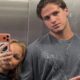 Mulher loira tira selfie com homem de camisa cinza em espelho de elevador