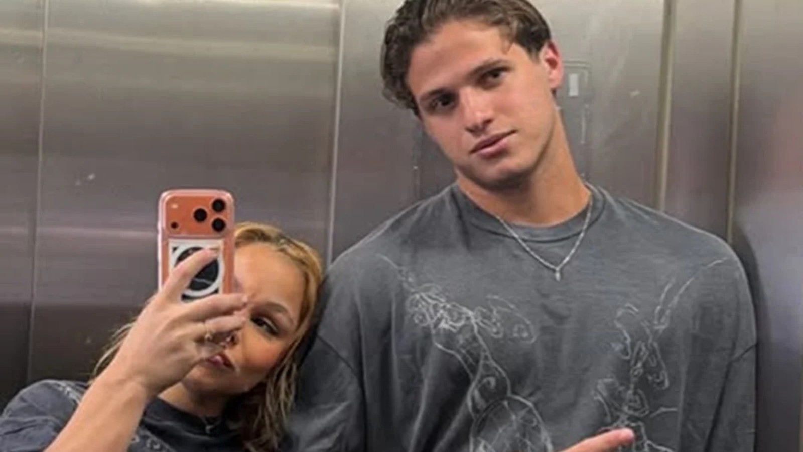 Mulher loira tira selfie com homem de camisa cinza em espelho de elevador