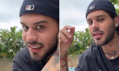 Homem de boné preto virado, barba, tatuagens no pescoço e braço, colares e brincos, sorrindo