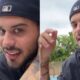 Homem de boné preto virado, barba, tatuagens no pescoço e braço, colares e brincos, sorrindo