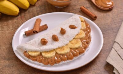 Tapioca recheada com banana fatiada e doce de leite, canela, em prato branco, com bananas ao fundo