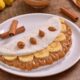 Tapioca recheada com banana fatiada e doce de leite, canela, em prato branco, com bananas ao fundo
