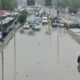 Rua alagada com carros, ônibus e pessoas sob céu nublado e chuva em Rio de Janeiro