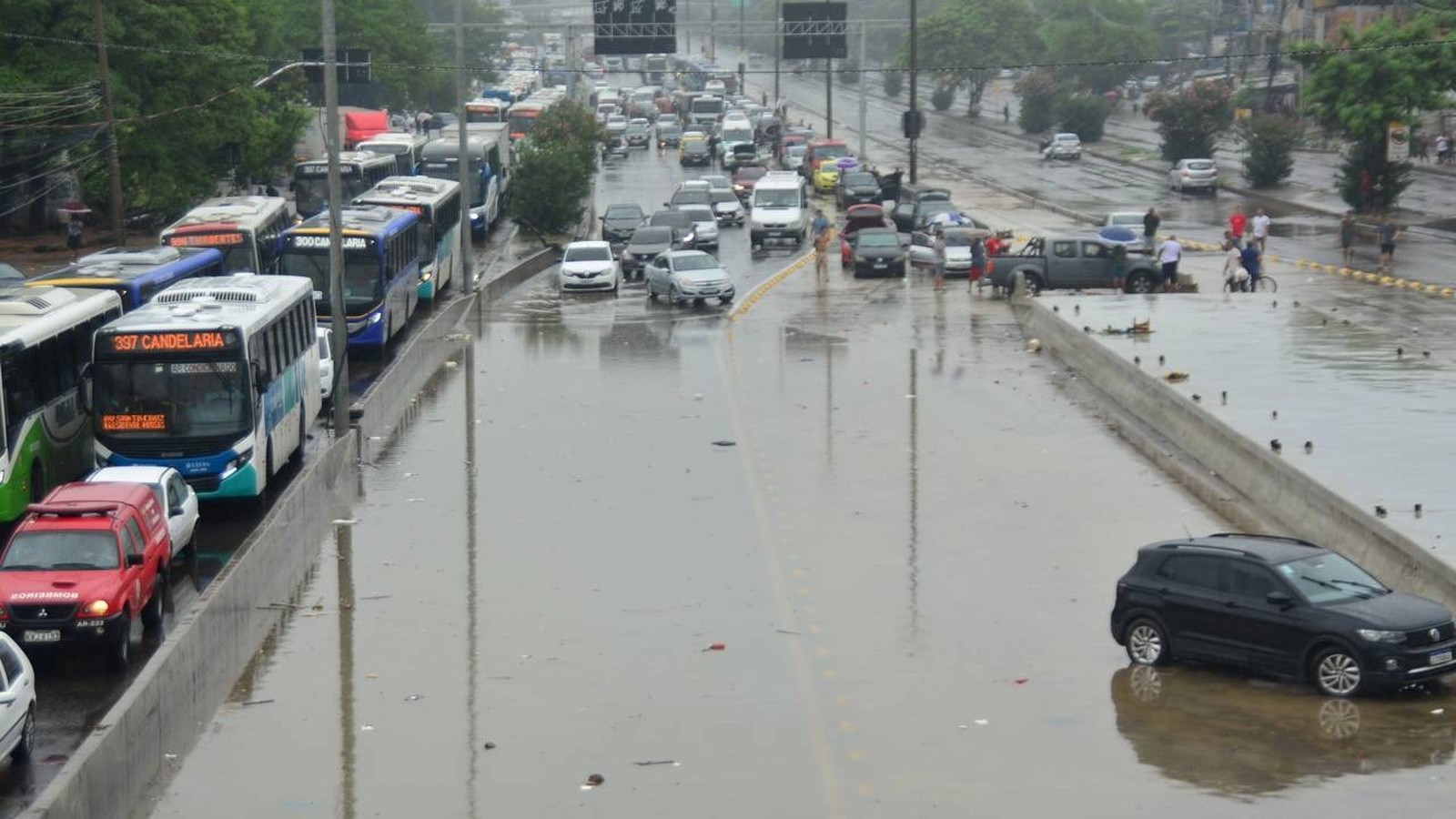 Rua alagada com carros, ônibus e pessoas sob céu nublado e chuva em Rio de Janeiro
