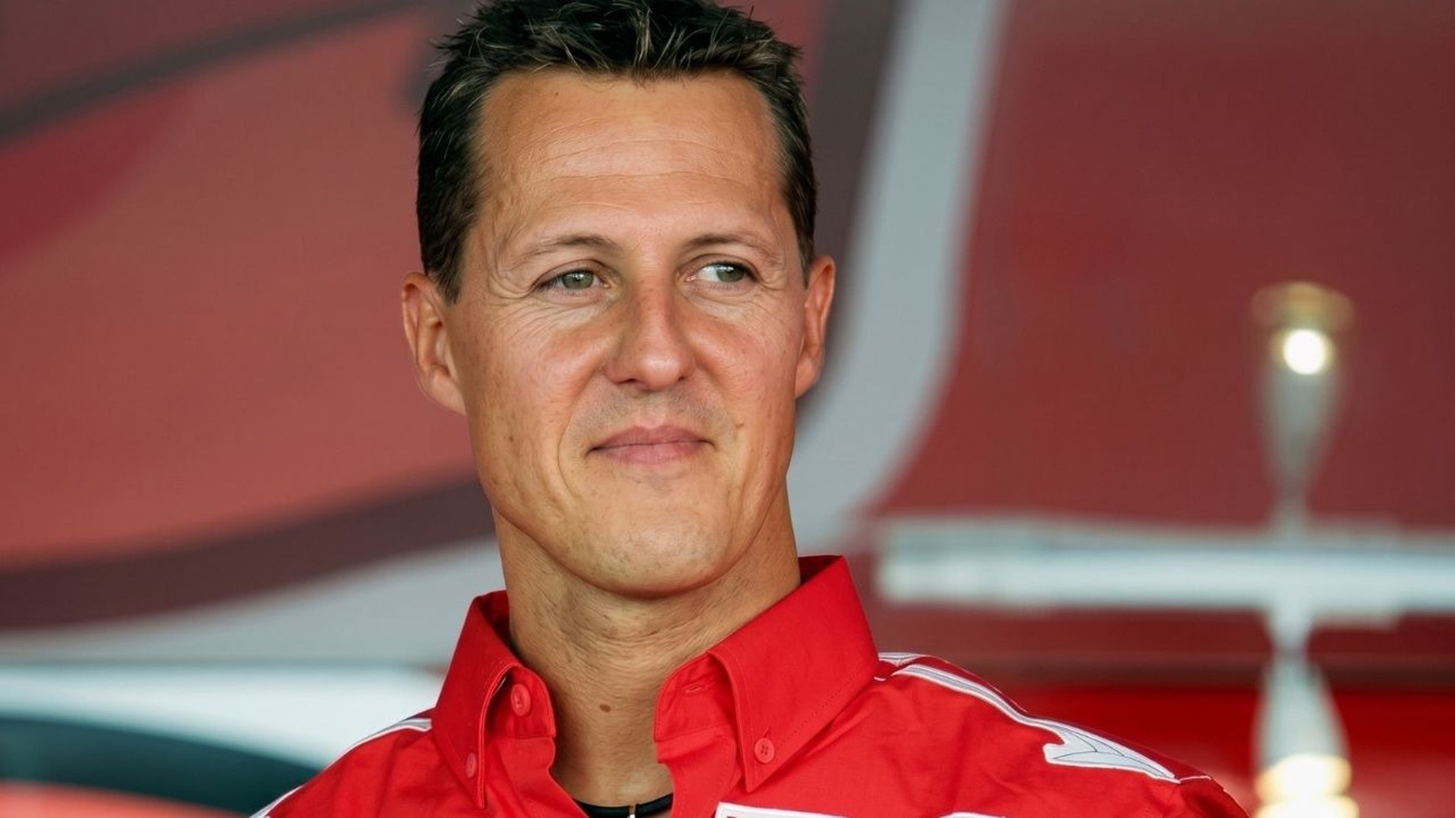 Close-up de Michael Schumacher sorrindo e olhando para o lado, vestindo camisa vermelha com detalhes brancos