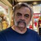 José Dumont, homem de cabelo e bigode grisalhos, vestindo camiseta azul, olhando para frente