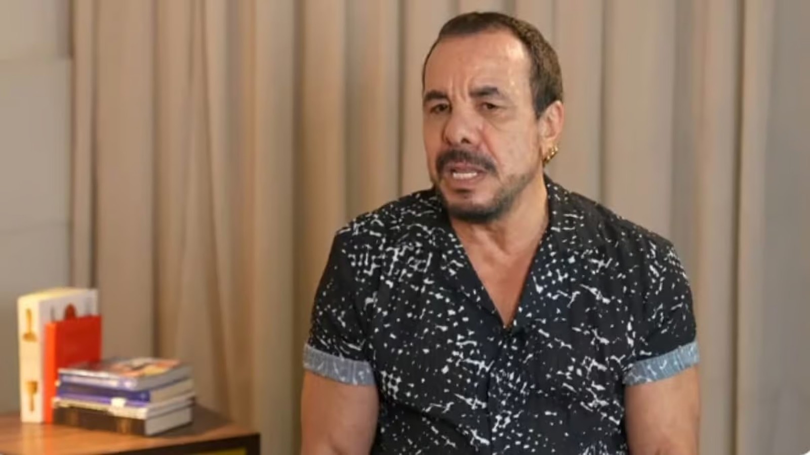 O cantor Ralf, com barba, camisa escura estampada e brinco dourado, falando.