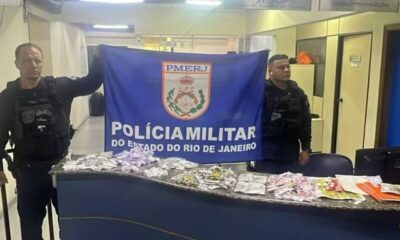 Dois policiais militares exibem banner da PMERJ e drogas apreendidas sobre uma mesa