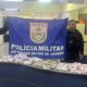 Dois policiais militares exibem banner da PMERJ e drogas apreendidas sobre uma mesa
