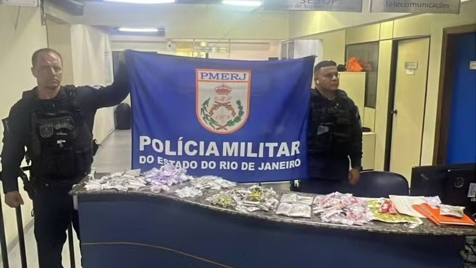 Dois policiais militares exibem banner da PMERJ e drogas apreendidas sobre uma mesa