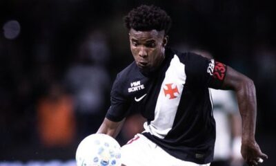 Jogador de futebol veste camisa preta e branca do Vasco com número 23 na manga, driblando bola.
