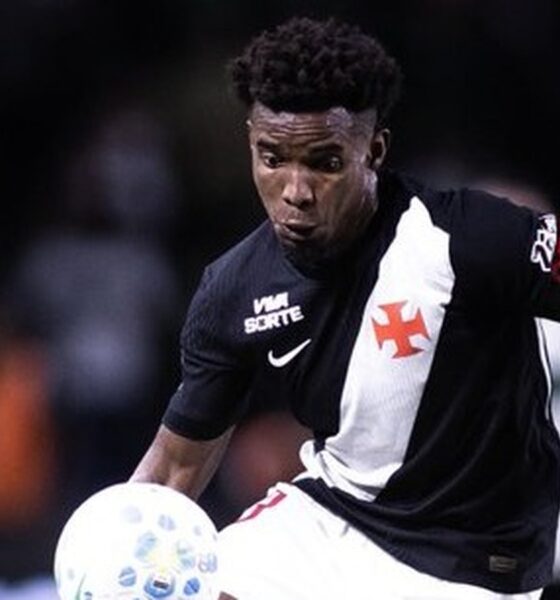 Jogador de futebol veste camisa preta e branca do Vasco com número 23 na manga, driblando bola.