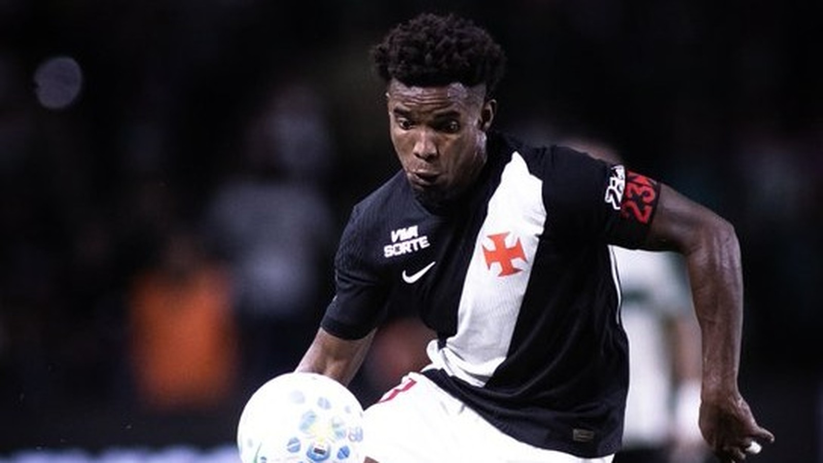 Jogador de futebol veste camisa preta e branca do Vasco com número 23 na manga, driblando bola.
