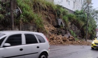 Rua parcialmente bloqueada por terra e pedras de deslizamento, com carros ao lado
