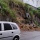 Rua parcialmente bloqueada por terra e pedras de deslizamento, com carros ao lado