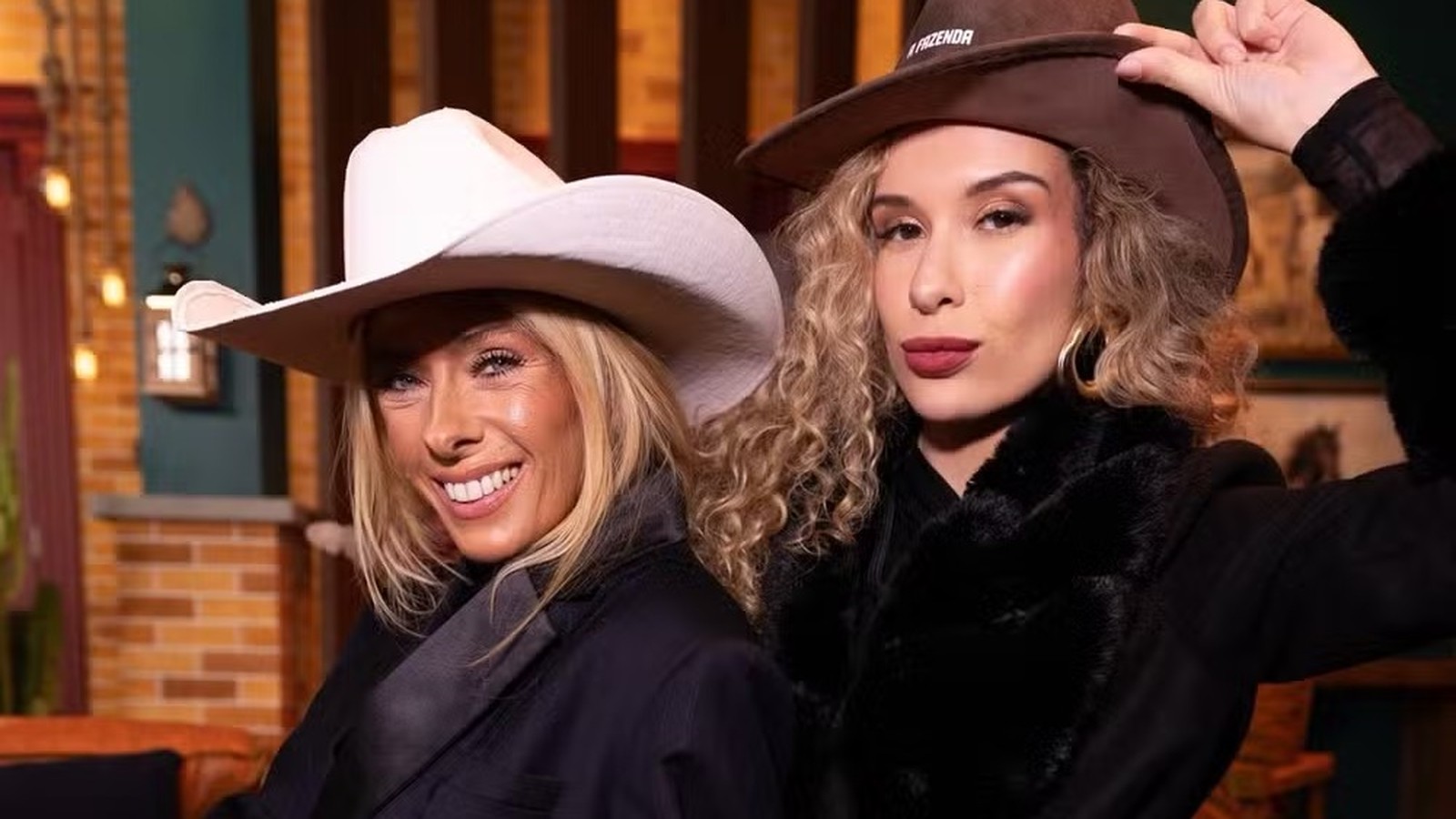 Duas mulheres de chapéus de cowboy. Uma loira sorrindo e outra com chapéu "A FAZENDA"