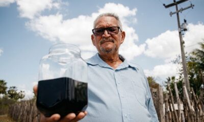 Homem idoso com óculos segura jarra de vidro com petróleo preto sob céu azul e nuvens