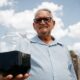 Homem idoso com óculos segura jarra de vidro com petróleo preto sob céu azul e nuvens