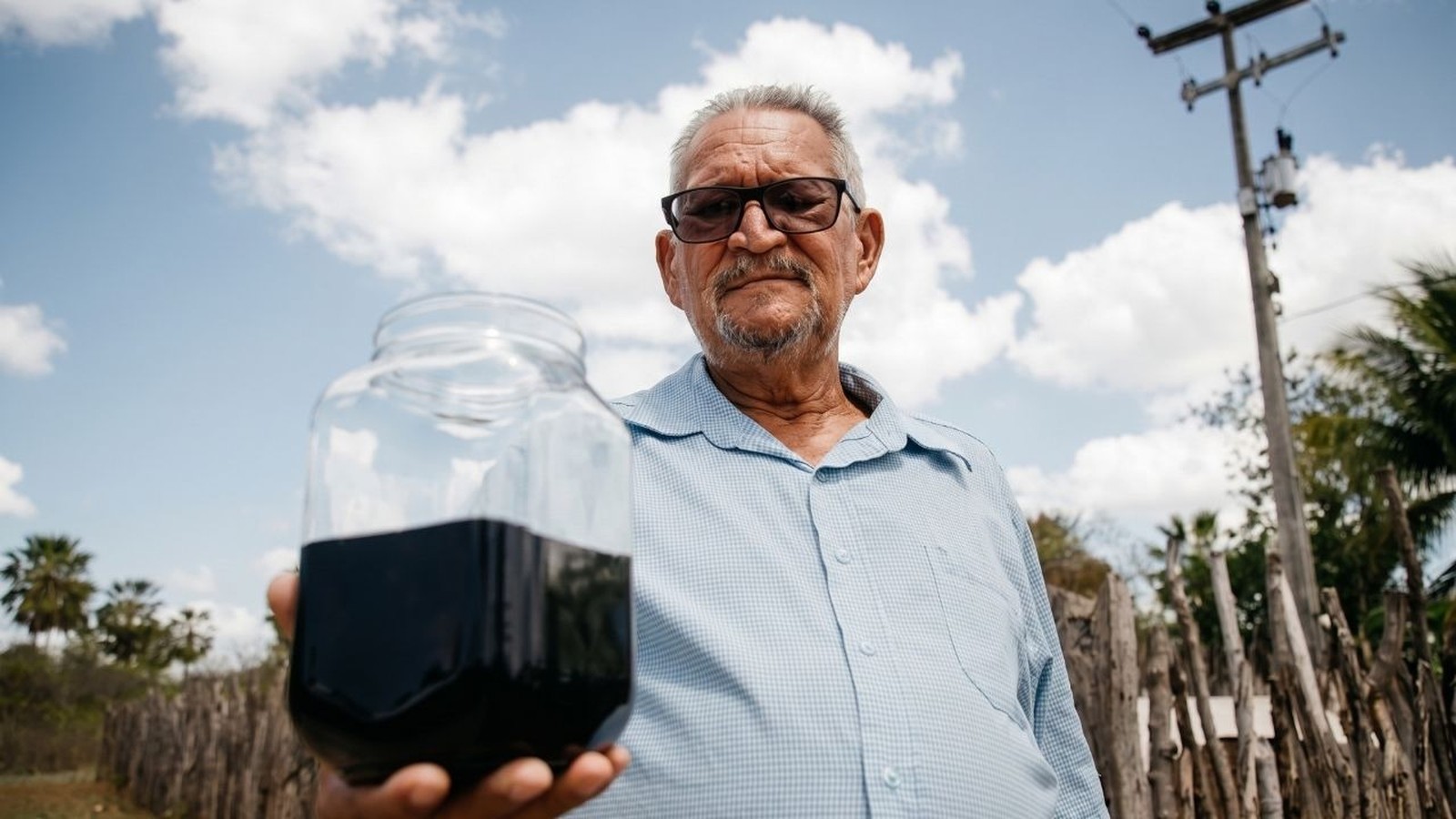 Homem idoso com óculos segura jarra de vidro com petróleo preto sob céu azul e nuvens