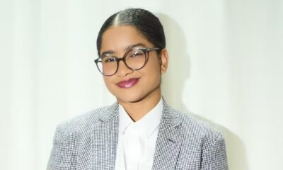 Close-up de mulher sorrindo, óculos redondos, batom vinho, camisa branca, blazer xadrez fundo claro