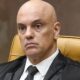 Homem careca em toga e terno escuros, gravata, sentado em cadeira de couro marrom, expressão séria