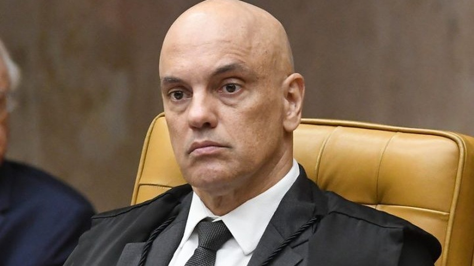 Homem careca em toga e terno escuros, gravata, sentado em cadeira de couro marrom, expressão séria