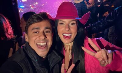 Selfie: Ana Castela sorrindo com homem em evento. Ela usa chapéu rosa e faz sinal