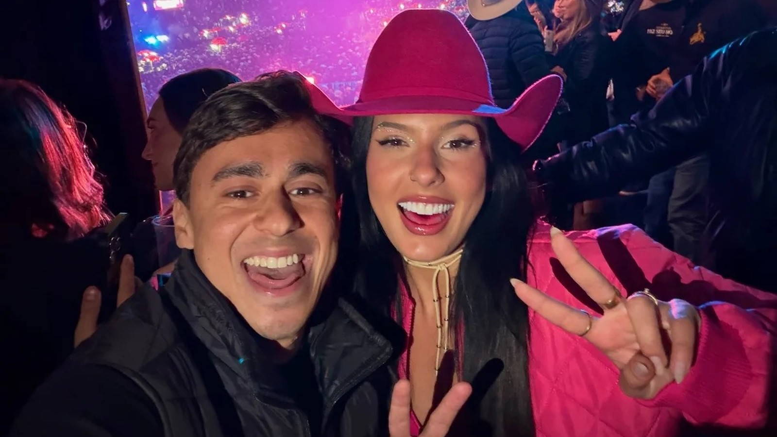 Selfie: Ana Castela sorrindo com homem em evento. Ela usa chapéu rosa e faz sinal