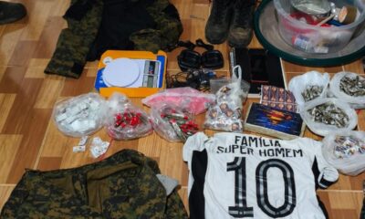 Várias sacolas de drogas, fardamento camuflado, balança, rádios, celular e camisa de time sobre piso