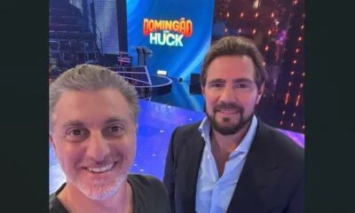 Luciano Huck e Daniel Vorcaro sorrindo no set do programa Domingão com Huck com telão ao fundo