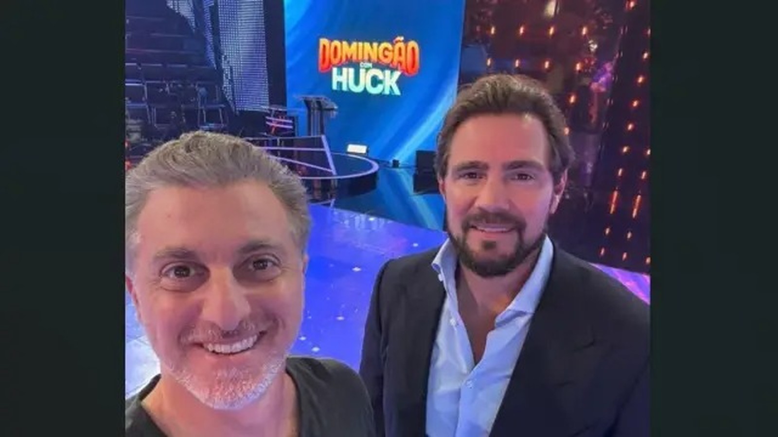 Luciano Huck e Daniel Vorcaro sorrindo no set do programa Domingão com Huck com telão ao fundo