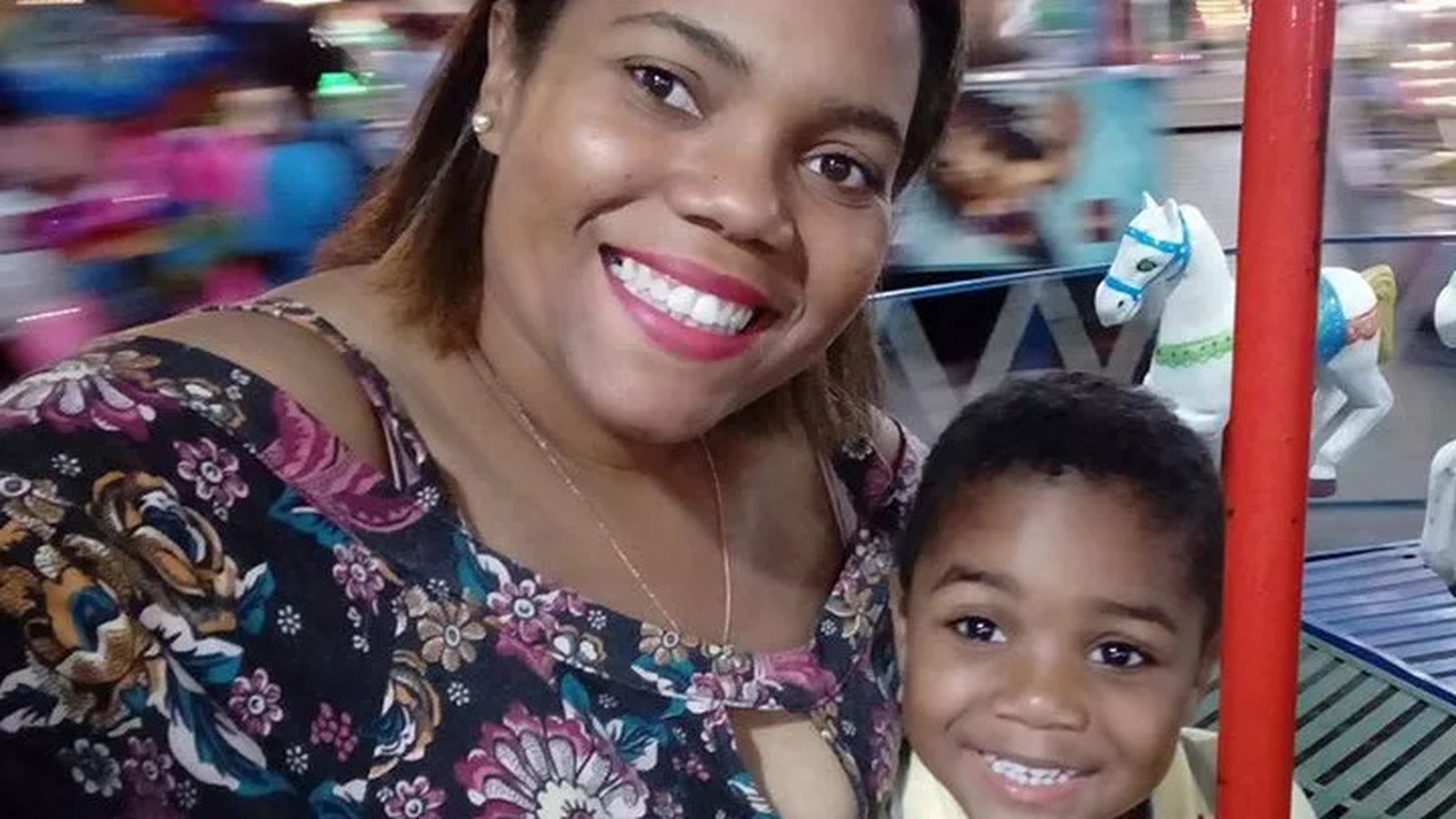 Mulher sorridente e menino em carrossel, com cavalo branco ao fundo