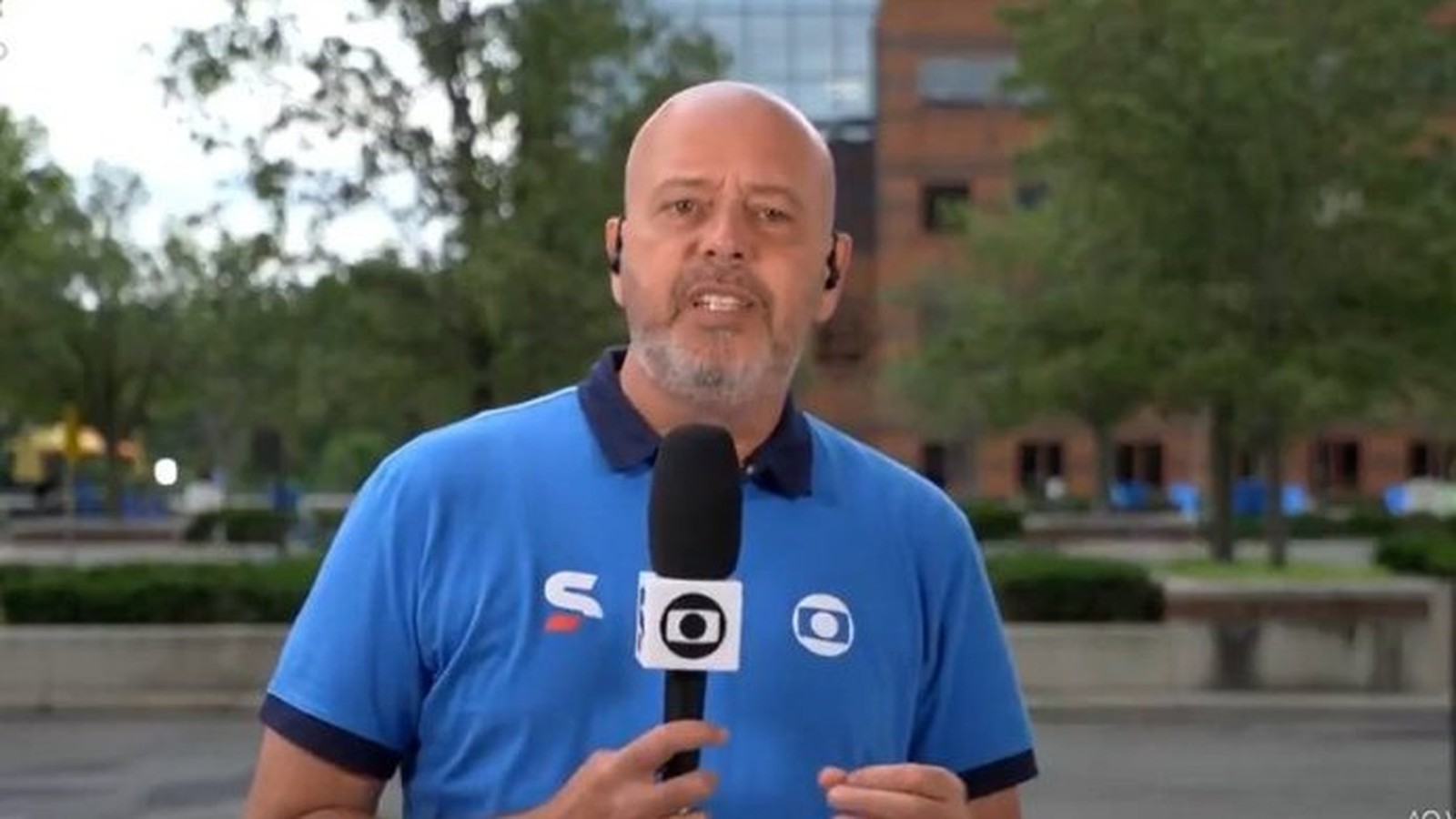 Jornalista Alex Escobar, careca e barbudo, em camisa polo azul segurando microfone da Globo