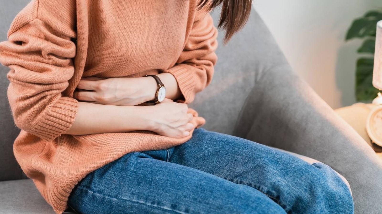 Mulher de suéter pêssego e jeans sentada, com as mãos no abdômen, em aparente desconforto ou dor.