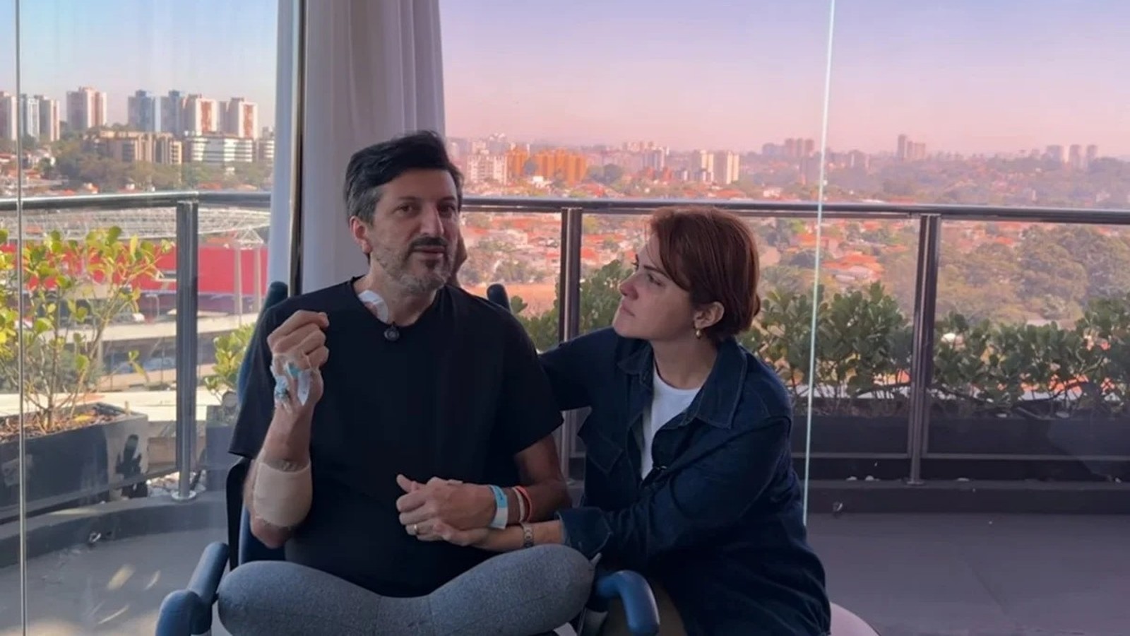 Homem com tubos médicos sentado com uma mulher, de mãos dadas, com vista para a cidade