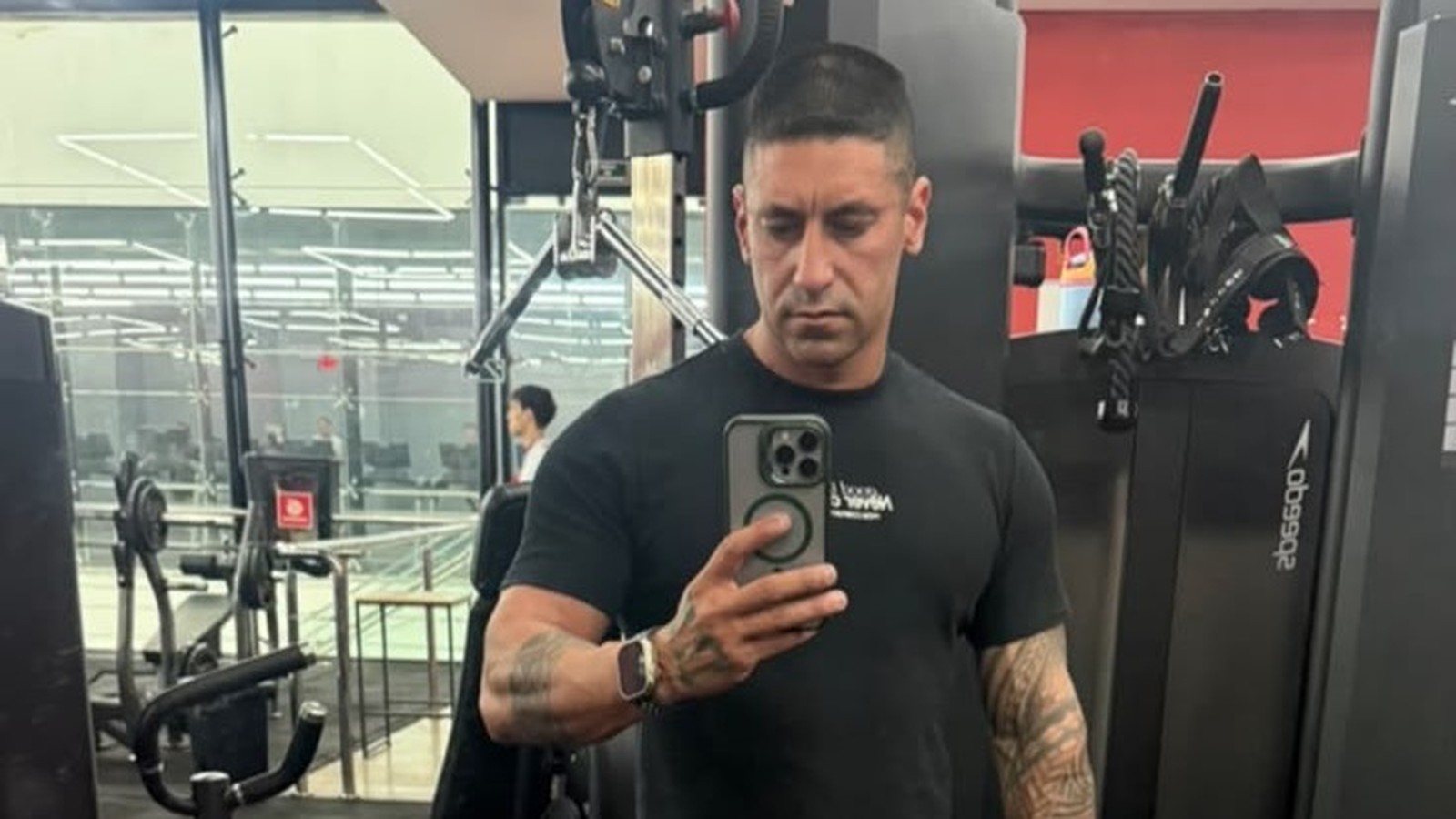 Homem com camiseta preta e tatuagens no braço tirando selfie em uma academia