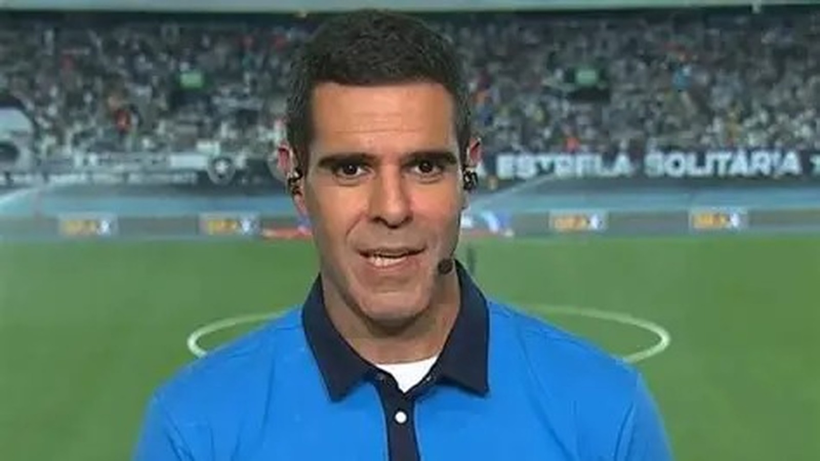 Homem de camisa polo azul e fones, com microfone, em campo de futebol com 'ESTRELA SOLITÁRIA'