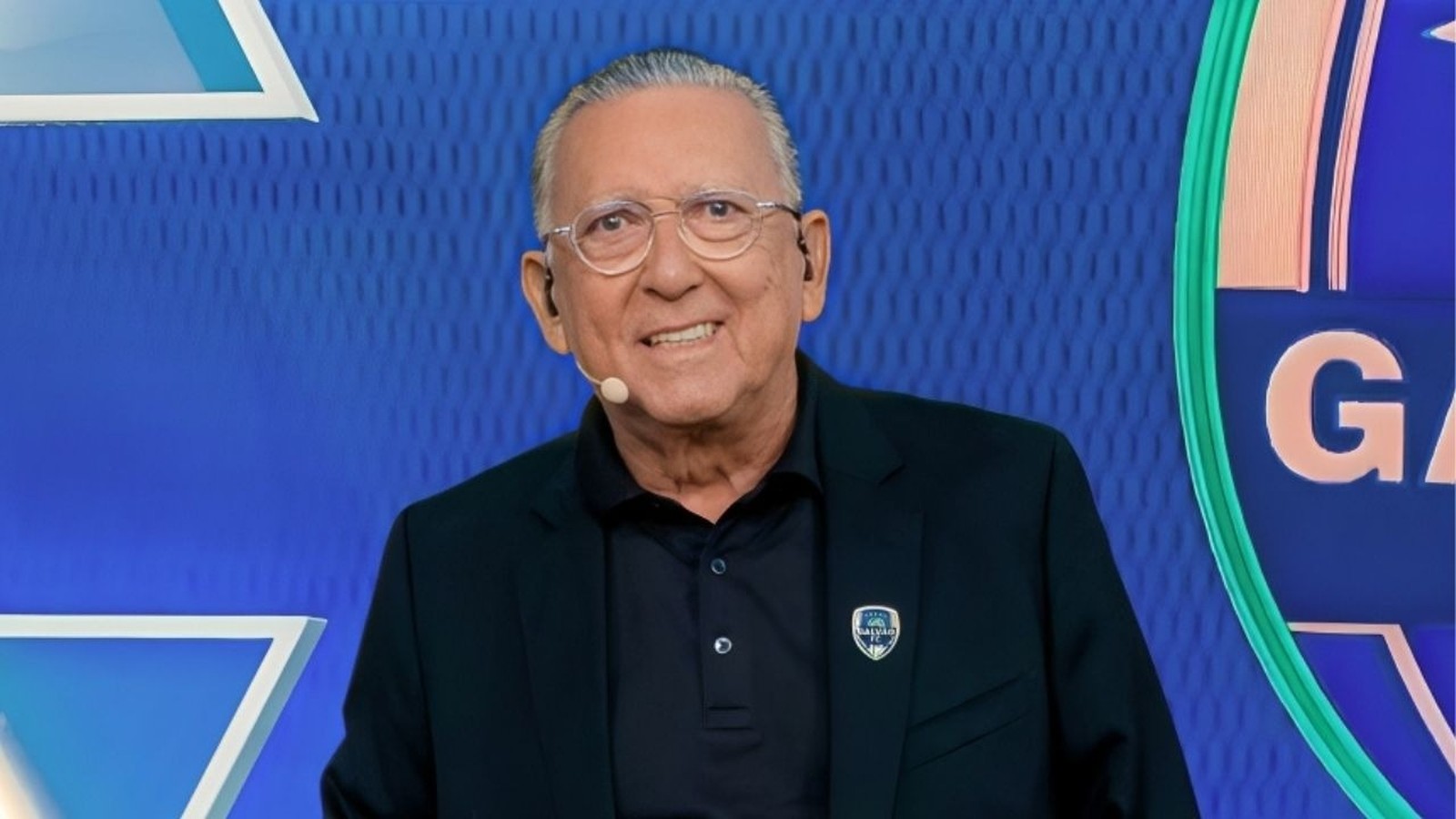 Galvão Bueno sorrindo, usando óculos e blazer, com broche 'Galvão FC' no estúdio
