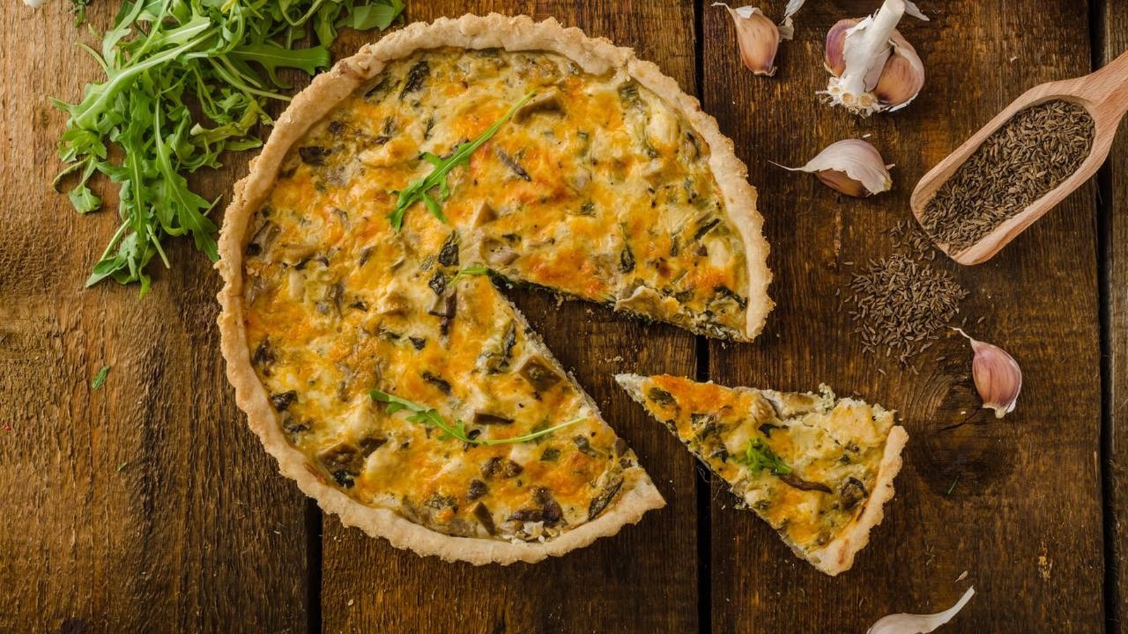 Quiche de alho-poró fatiada em mesa de madeira rústica, com alho e sementes de cominho