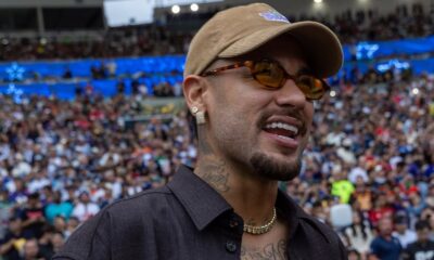 Close-up de Neymar sorrindo em estádio lotado, usando boné, óculos e corrente