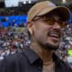 Close-up de Neymar sorrindo em estádio lotado, usando boné, óculos e corrente